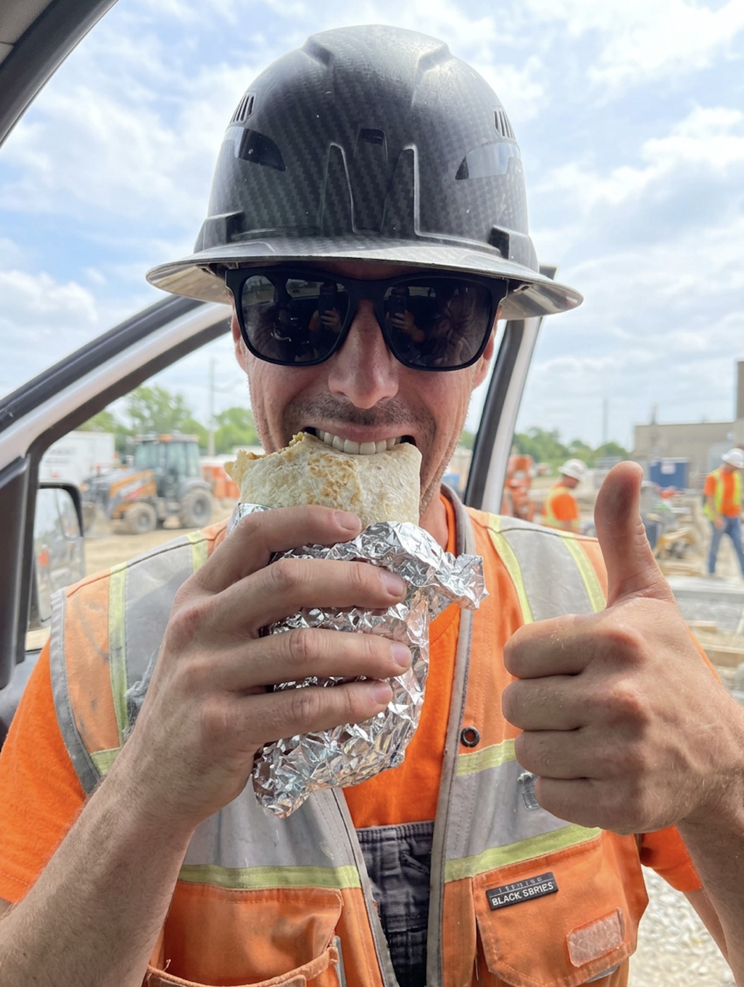 Kevin Foxcroft in a hard hat, sunglasses, and an orange hi-vis vest, biting into a foil-wrapped burrito and giving a thumbs up on a job site.